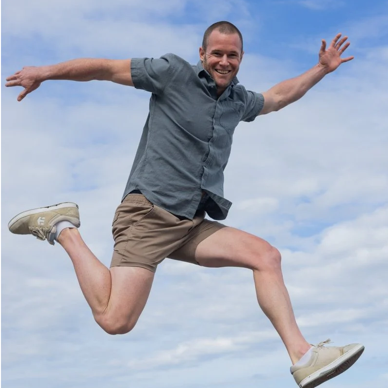 Man wearing light brown men's shorts made from soft, breathable fabric paired with a short-sleeve grey shirt, jumping joyfully outdoors against a cloudy sky.