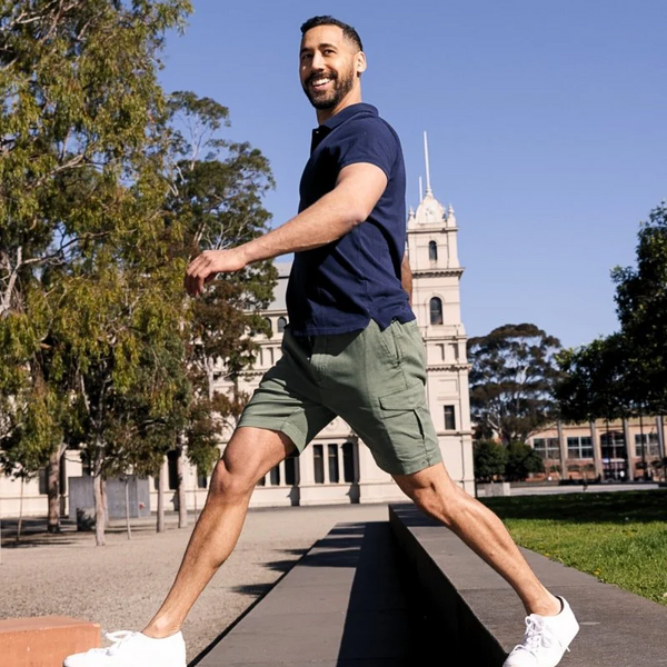 Man wearing olive green men's shorts made from soft, breathable fabric paired with a navy blue polo shirt, walking outdoors on a sunny day.