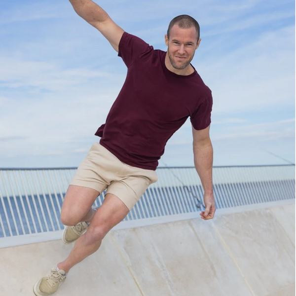 Man wearing beige men's shorts made from lightweight, breathable fabric paired with a burgundy t-shirt, balancing outdoors against a clear sky.