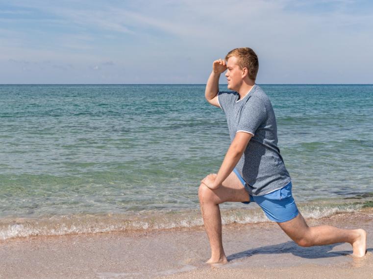 A young man does a lunge on a sandy beach while looking out at the ocean, holding his hand above his eyes to shield from the sun. He wears a gray t-shirt and blue frankster shorts. The sea appears calm under a partly cloudy sky.