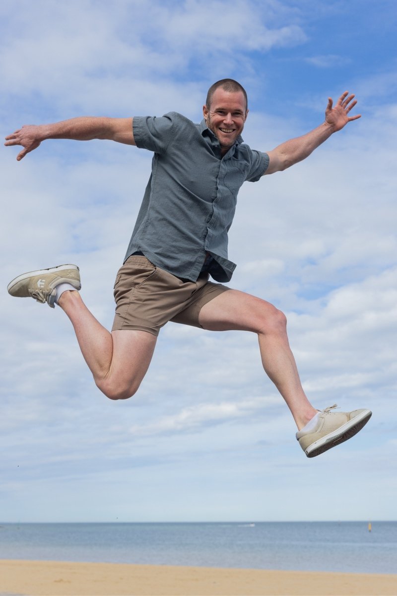 Man jumping on beach wearing Nomads * The Shorties dark khaki stretch cotton men's shorts by Frankster