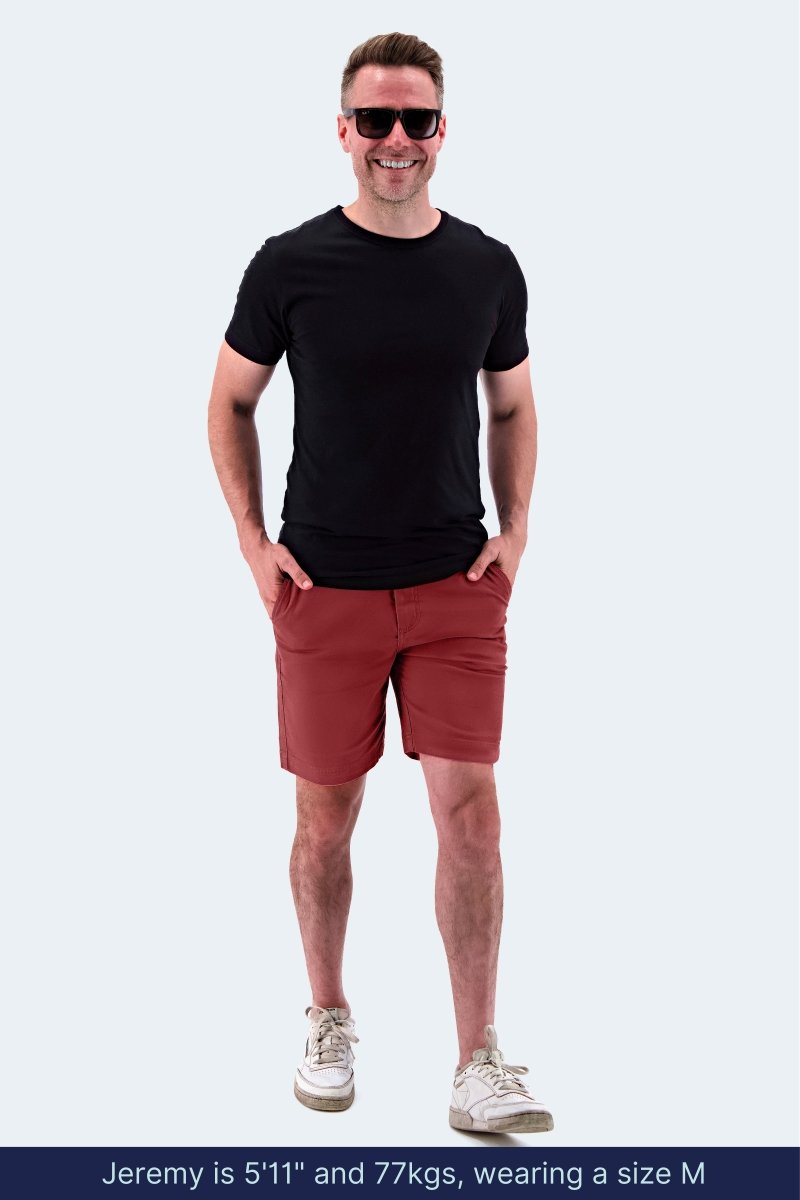 Man wearing Frankster Pioneers * The Everydays burgundy stretch cotton men's shorts with black t-shirt and white sneakers walking outdoors