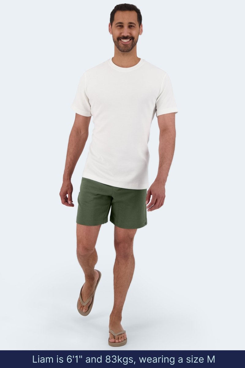 Man wearing Frankster Rebels * The Comfies olive green cotton men's shorts and white t-shirt walking indoors