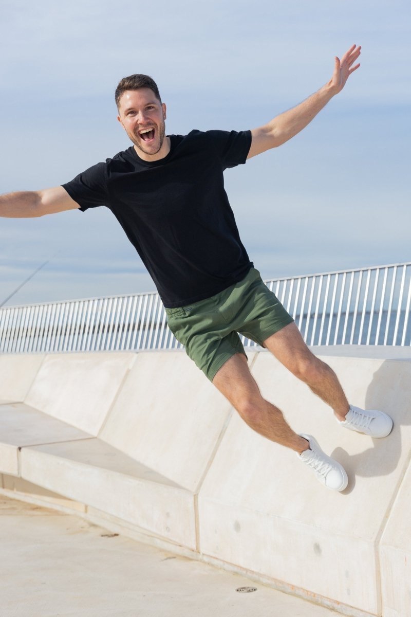 Man wearing Frankster Rebels * The Shorties army green stretch cotton men's shorts with a black t-shirt and white sneakers outdoors