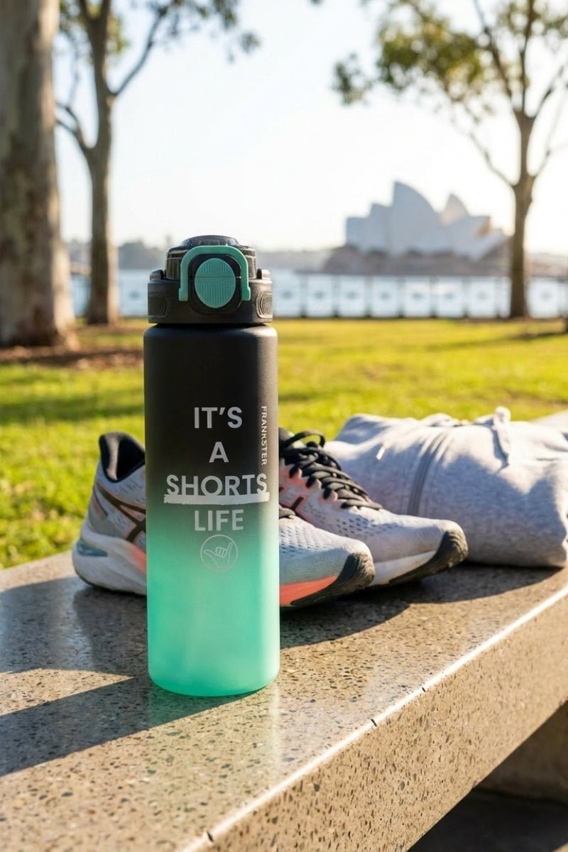 Frankster drinkware bottle in gradient bottle green and black placed outdoors with casual men's shorts and running shoes, promoting active lifestyle