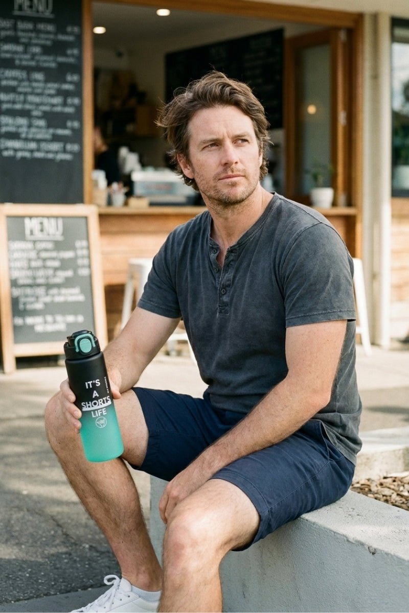 Man wearing Frankster Shorts Life Bottle Green Black men's shorts in a casual dark wash sitting outdoors holding a matching green and black bottle, showcasing comfortable men's shorts for everyday wear.