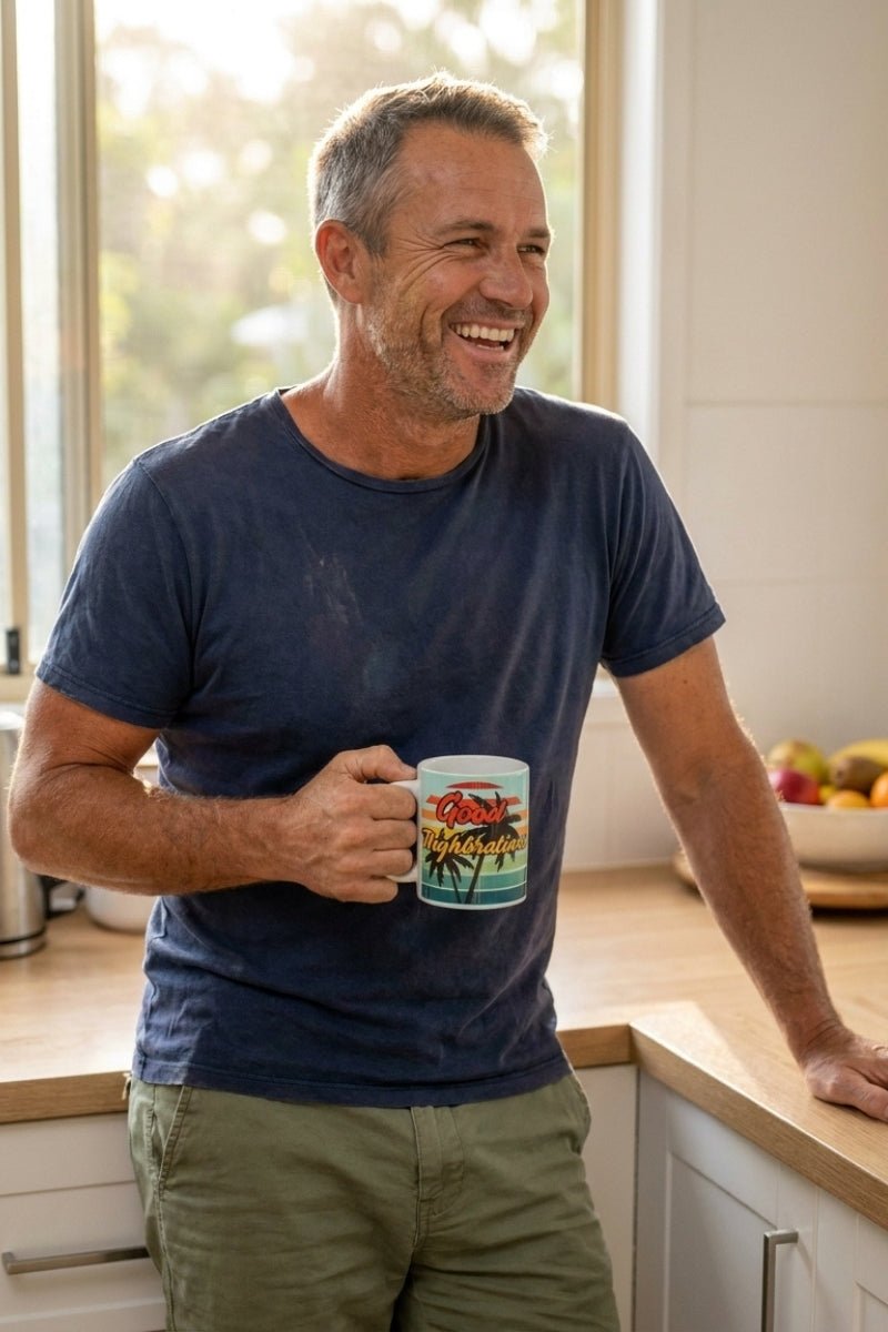 Man wearing casual olive-green men's shorts and a worn navy-blue t-shirt, holding The Good Thighbrations Mug in a bright kitchen setting, showcasing relaxed comfort linked to Frankster men's shorts.