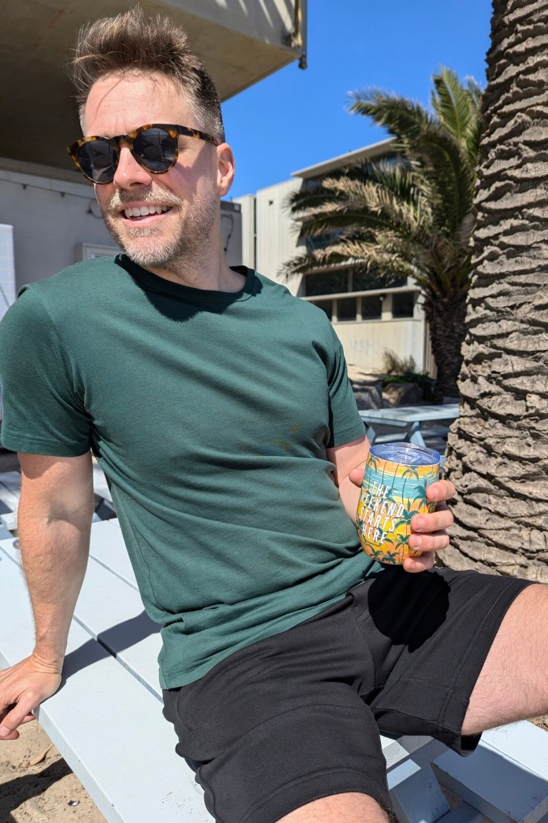 Man wearing black cotton men's shorts and a green t-shirt holding The Tropical Weekend Tumbler by Frankster outdoors