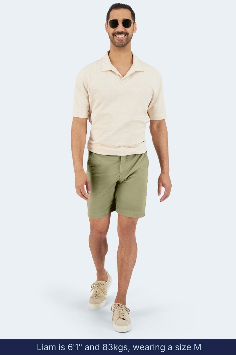 Man wearing Frankster Virgins * The Everydays olive green stretch cotton men's shorts with beige shirt and sneakers walking outdoors