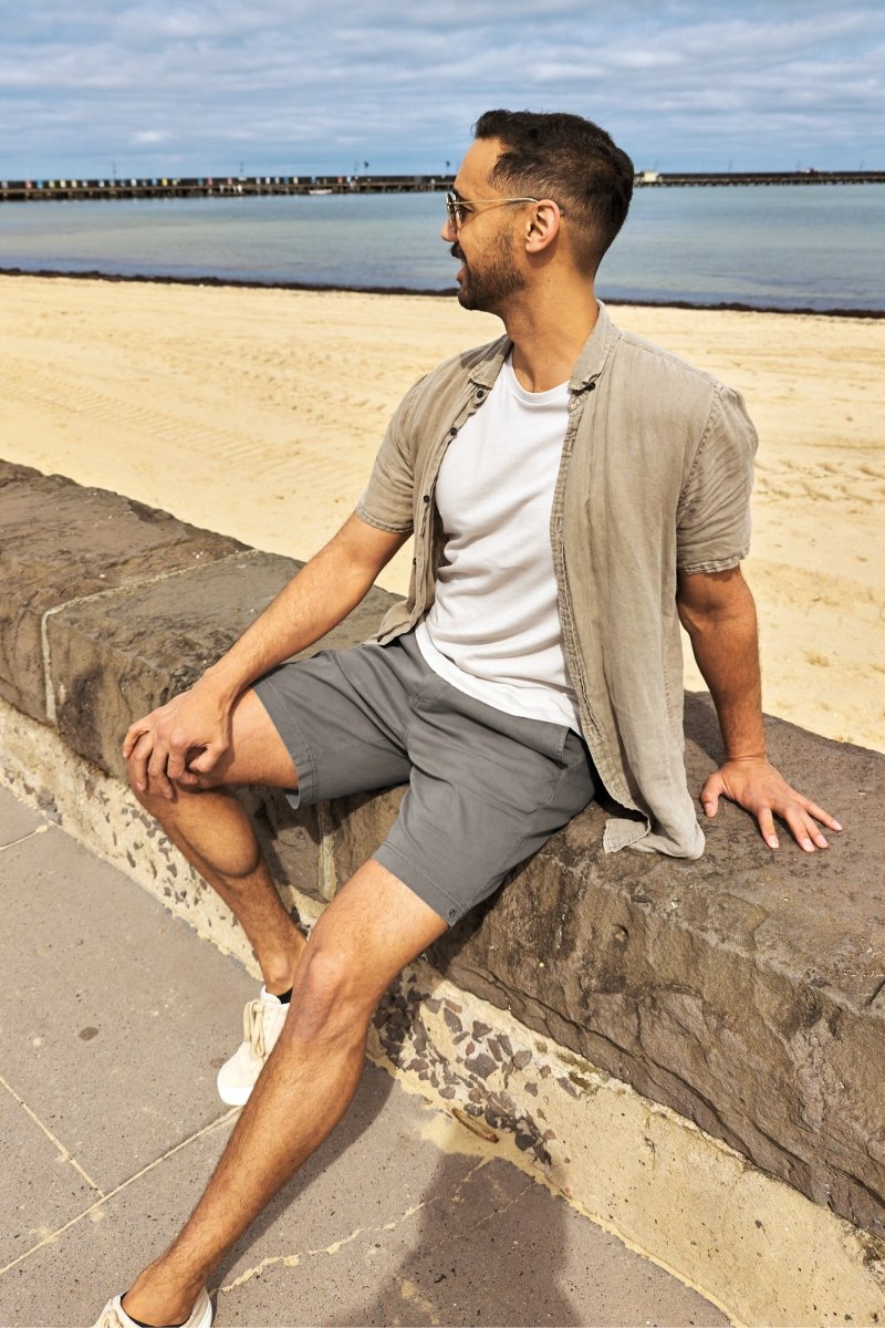 Man wearing Volcans * The Everydays dark grey Frankster men's shorts sitting by the beach in casual shirt and white tee
