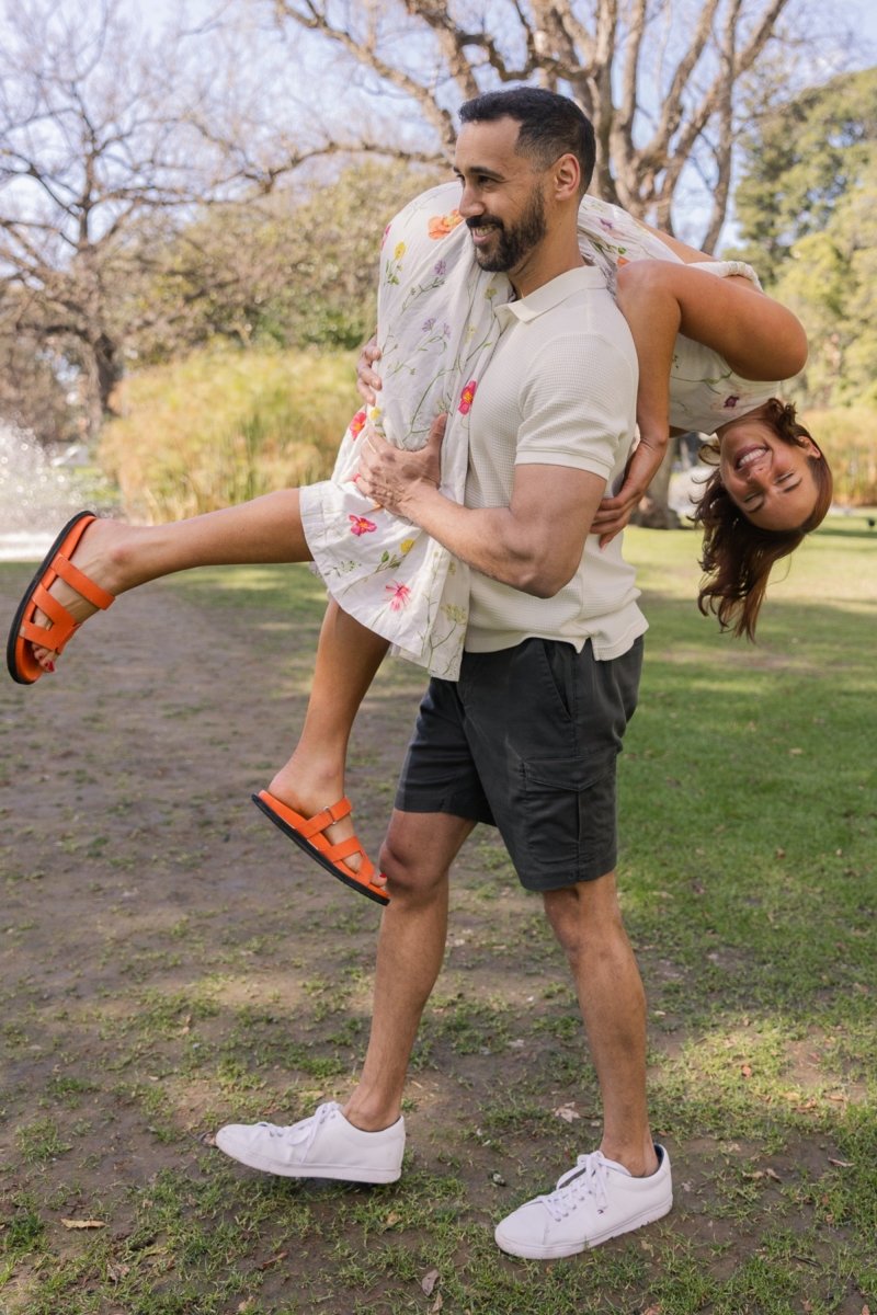 Man wearing dark grey Volcans * The Aussies Frankster men's shorts outdoors carrying woman in floral dress and orange sandals