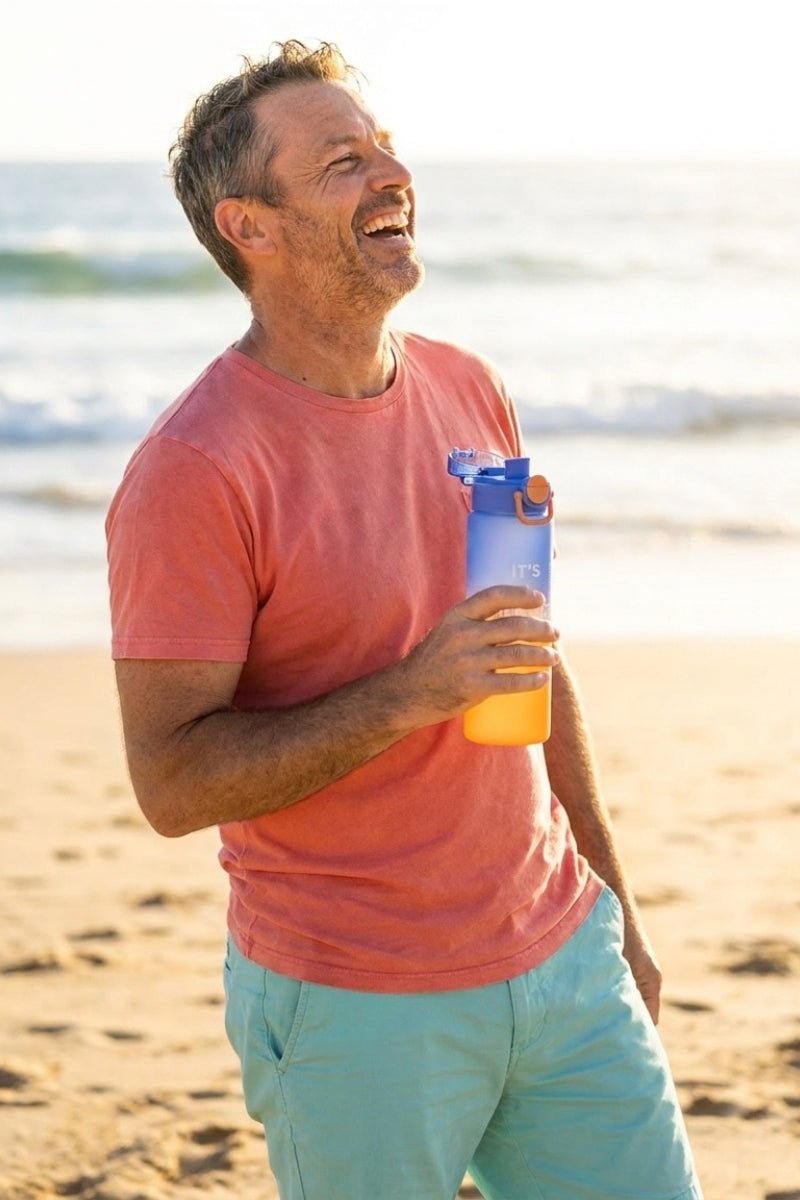 Man smiling on the beach wearing light teal men's shorts paired with a soft orange t-shirt, holding a Water Bottle Orange Blue by Frankster, showcasing casual comfort and style.