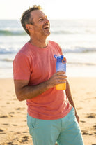 Man smiling on the beach wearing light teal men's shorts paired with a soft orange t-shirt, holding a Water Bottle Orange Blue by Frankster, showcasing casual comfort and style.