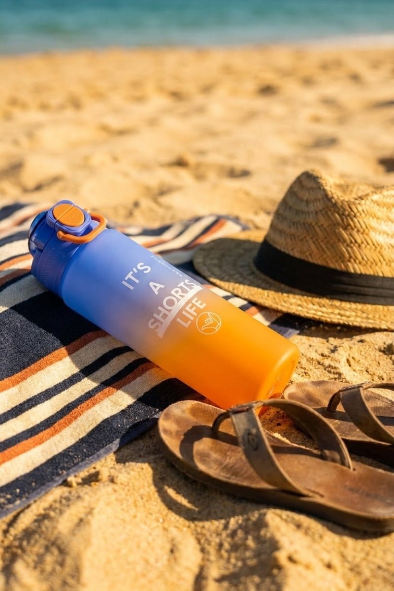 Water Bottle Orange Blue resting on a striped towel beside a straw hat and worn sandals on sandy beach, evoking casual men's shorts day.